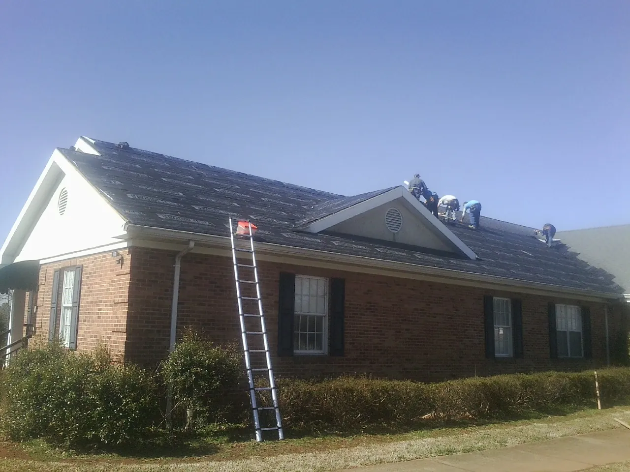 Crew tearing shingles off a brick ranch roof with a ladder set — Emergency Roof Repair work by our crew in Wendell